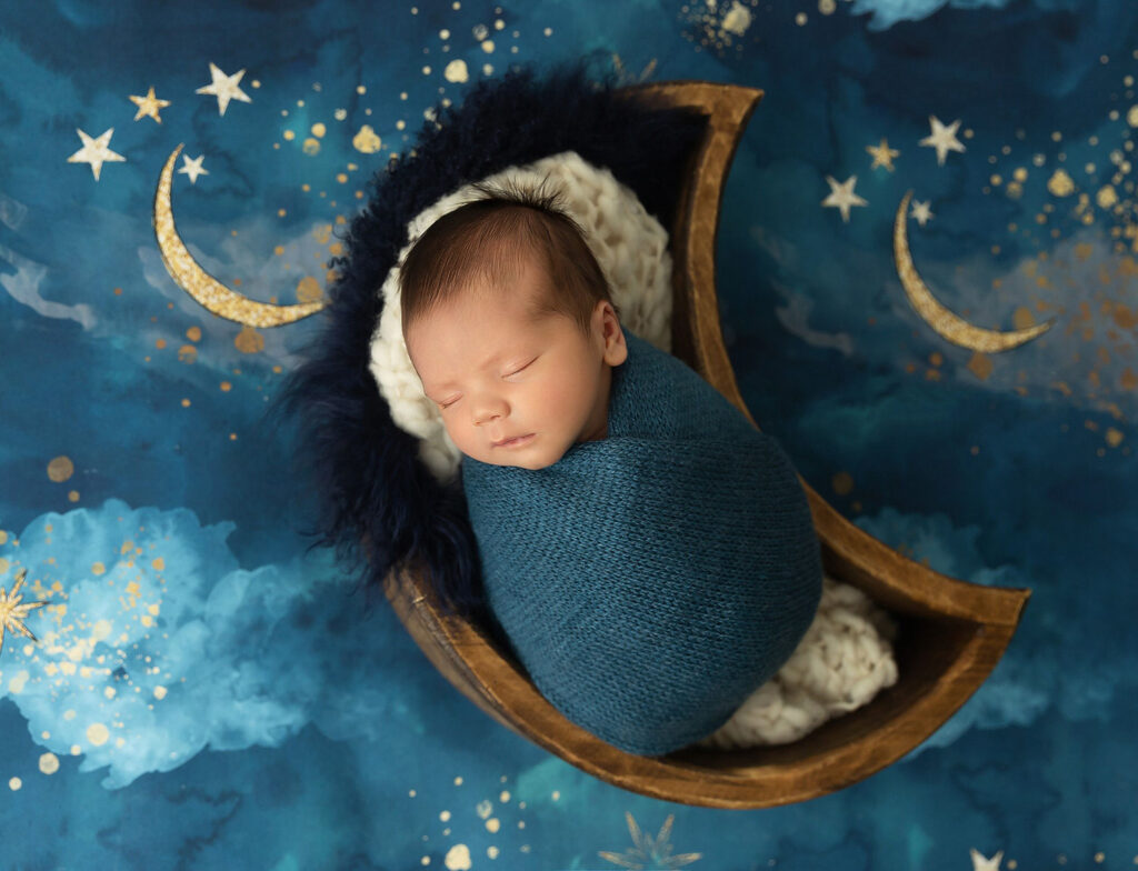 Newborn baby boy swaddled in a moon prop on a starry background